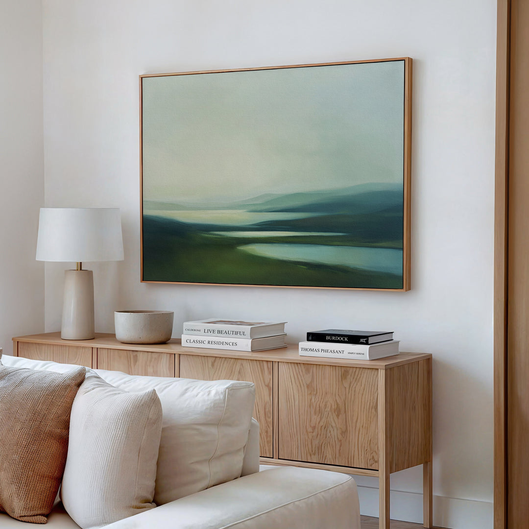 A modern living room featuring a light wood sideboard, white lamp, books, bowl, and Gentle Valleys Canvas Art – Horizontal above a cream-colored sofa for serene and stylish decor.