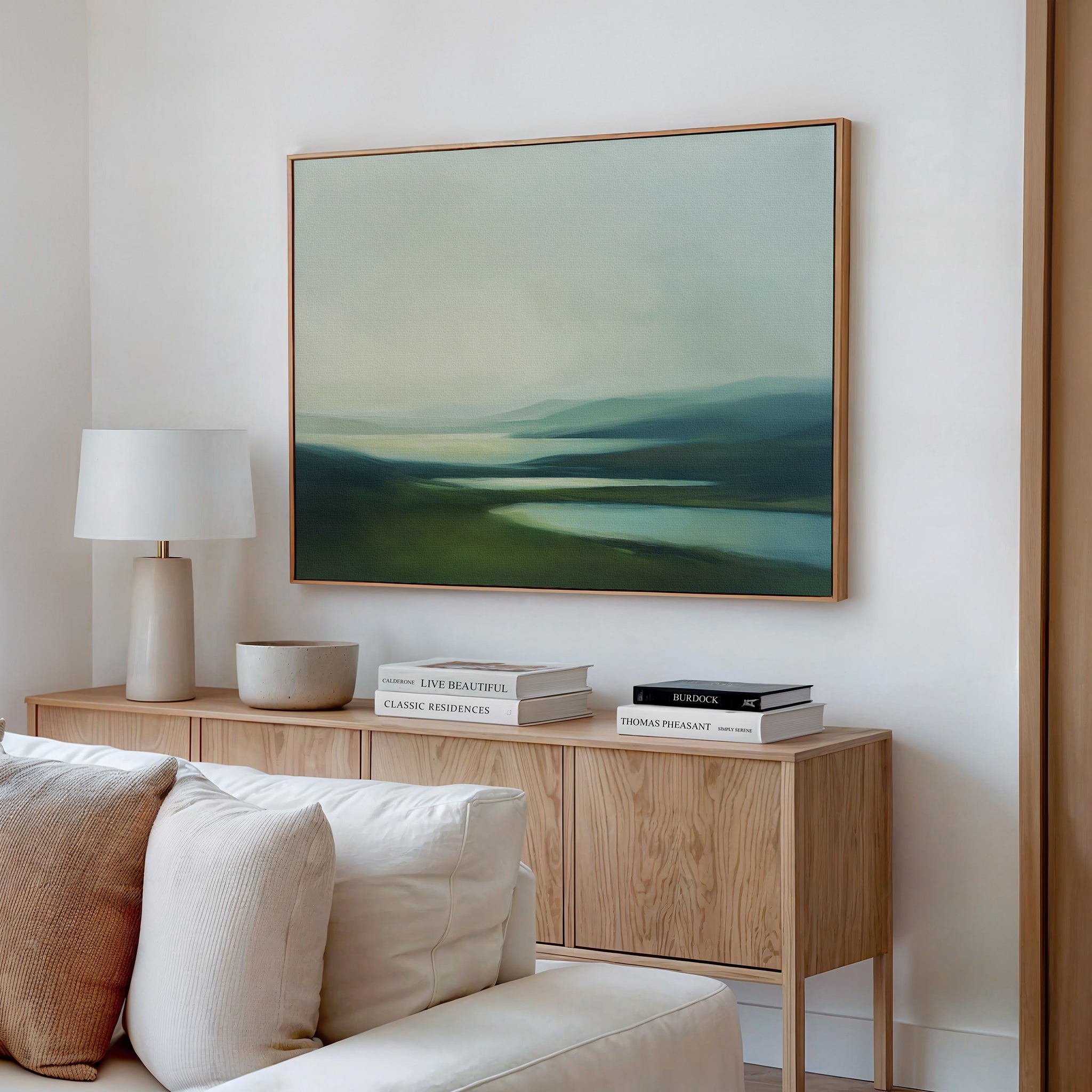 A modern living room featuring a light wood sideboard, white lamp, books, bowl, and Gentle Valleys Canvas Art – Horizontal above a cream-colored sofa for serene and stylish decor.