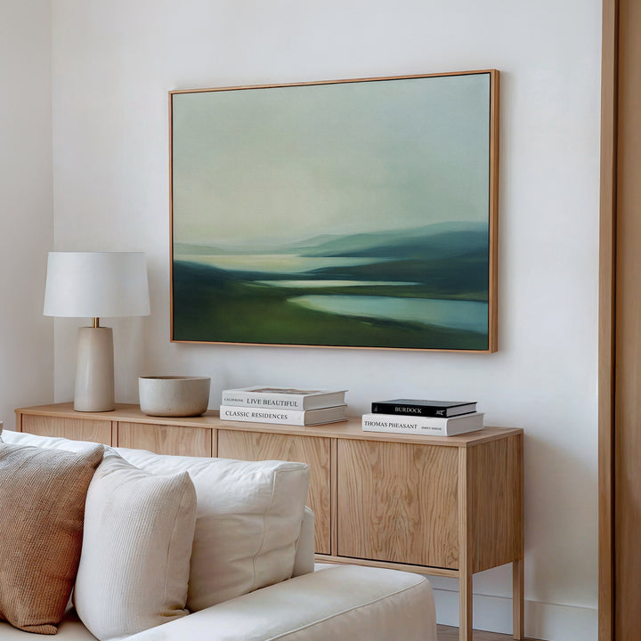 A modern living room featuring a light wood sideboard, white lamp, books, bowl, and Gentle Valleys Canvas Art – Horizontal above a cream-colored sofa for serene and stylish decor.