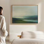A person in a white shirt stands in a minimalist room, admiring the Gentle Valleys Canvas Art – Horizontal above a light-colored sofa. This framed artwork features tranquil hills and water in soft, muted tones, enhancing the serene living room decor.