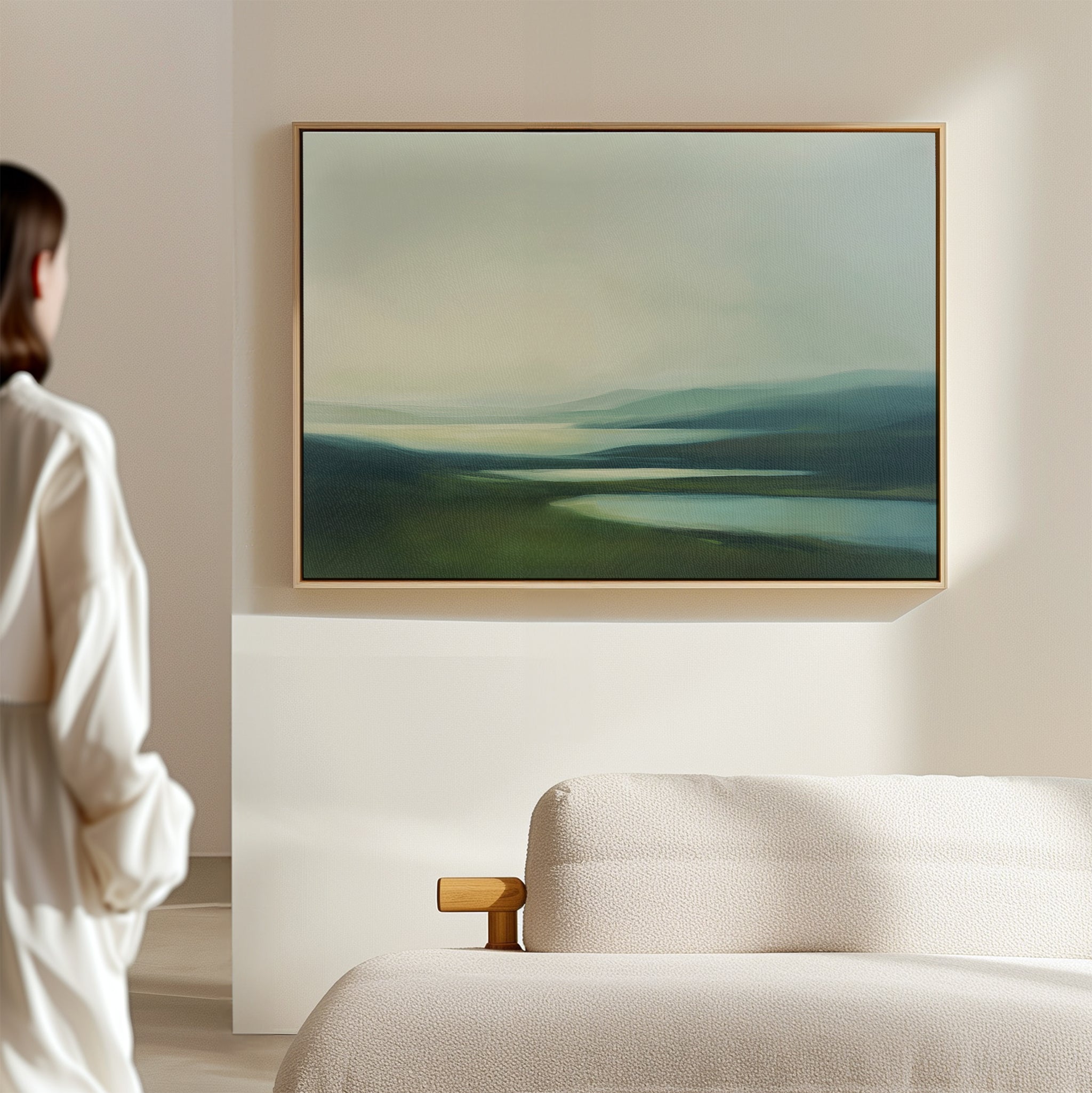 A person in a white shirt stands in a minimalist room, admiring the Gentle Valleys Canvas Art – Horizontal above a light-colored sofa. This framed artwork features tranquil hills and water in soft, muted tones, enhancing the serene living room decor.