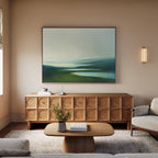 A modern living room with neutral tones features a wooden sideboard, the Gentle Valleys Canvas Art – Horizontal as serene wall decor, a low coffee table with books, a light beige sofa on a soft rug, and a wall sconce providing soft lighting.
