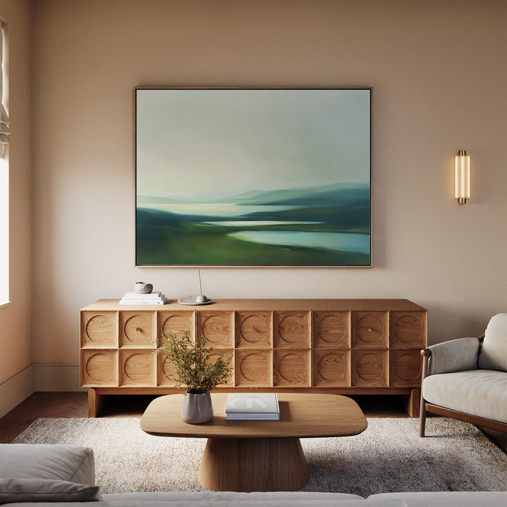 A modern living room with neutral tones features a wooden sideboard, the Gentle Valleys Canvas Art – Horizontal as serene wall decor, a low coffee table with books, a light beige sofa on a soft rug, and a wall sconce providing soft lighting.