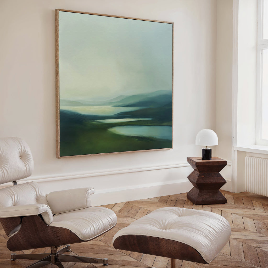 A modern, minimalist living room with a white lounge chair and ottoman, a geometric wooden side table with a small lamp, and Gentle Valleys Abstract Canvas Art featuring green hills and water—perfect for refined decor.