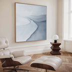 A modern living room with a white leather lounge chair and ottoman, wood flooring, a side table with a white lamp, and the Glacial Flow Canvas Art in soft blue and gray tones creates chic modern decor.
