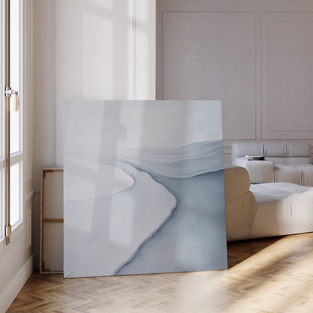 The Glacial Flow Canvas Art, featuring soft abstract patterns in white and gray, leans against the wall of a modern living room with parquet floors and white furniture—ideal for contemporary decor.