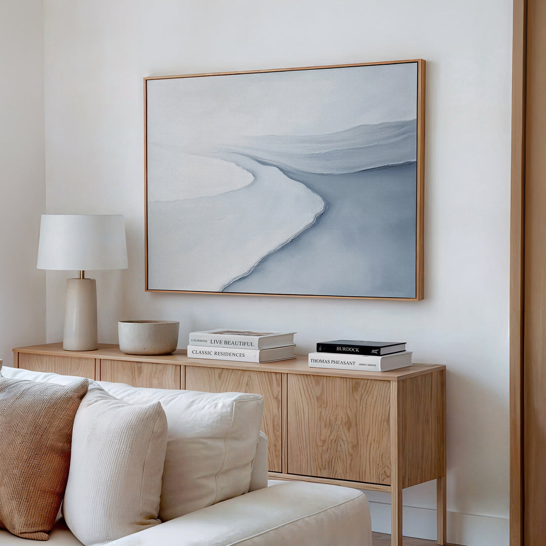 A minimal, modern living room features a light wood sideboard with books, a bowl, and a white lamp. Above, Glacial Flow Canvas Art – Horizontal in soft blue and gray hangs on the white wall. A beige couch anchors the decor.