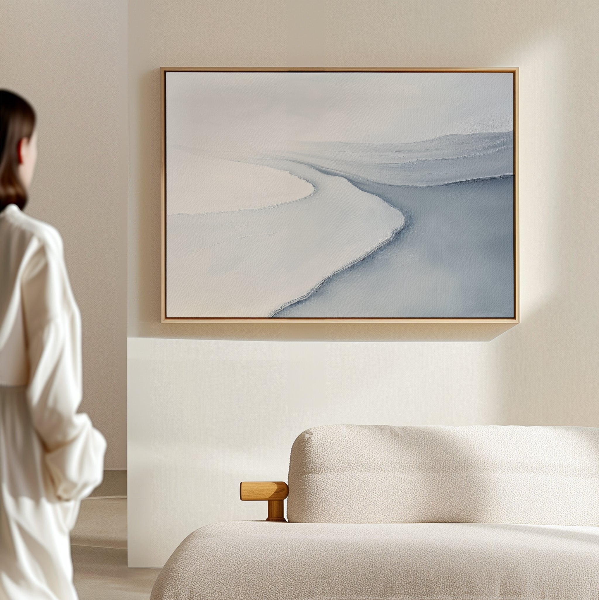 A person stands in a softly lit room, admiring the Glacial Flow Canvas Art – Horizontal with pale blue and white tones above a light-colored sofa—a perfect modern living room decor accent.