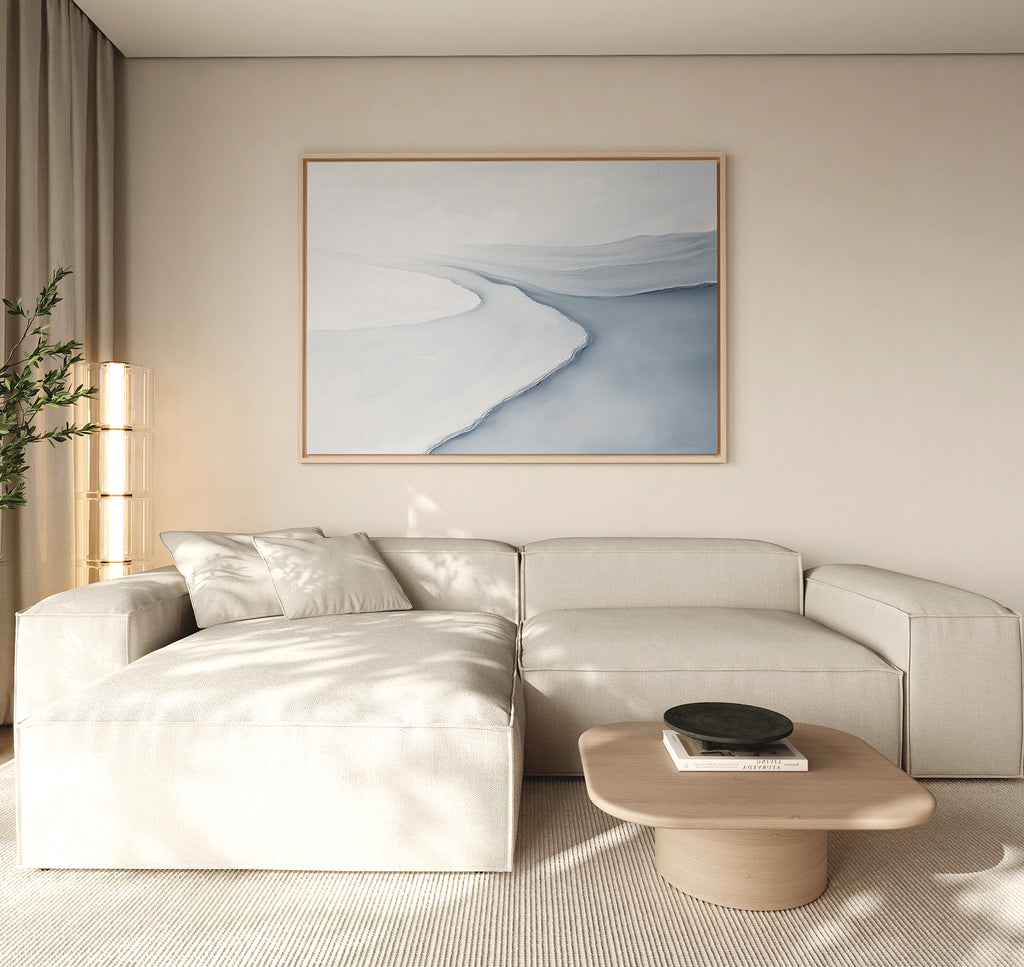 A modern living room features a light beige sectional sofa, neutral walls, a wooden coffee table, a black bowl, a tall plant, and the Glacial Flow Canvas Art – Horizontal in blue and white tones displayed above the sofa.