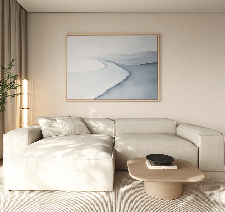 A modern living room features a light beige sectional sofa, neutral walls, a wooden coffee table, a black bowl, a tall plant, and the Glacial Flow Canvas Art – Horizontal in blue and white tones displayed above the sofa.