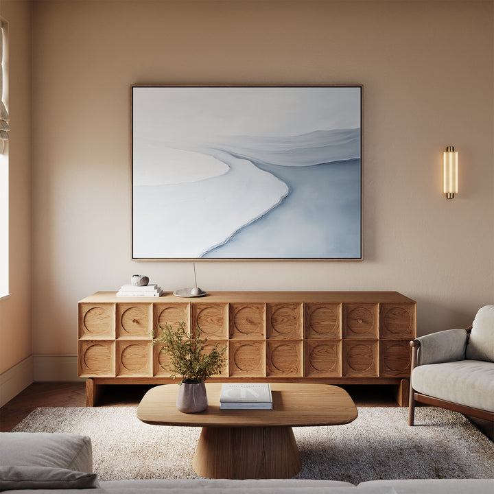 A modern living room showcases a light sofa, wooden coffee table, and sideboard. Above the sideboard hangs the Glacial Flow Canvas Art – Horizontal in blue and white, while a small vase with flowers sits on the coffee table.