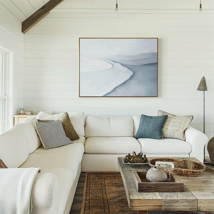 A cozy living room with a white sectional sofa, assorted pillows, a rustic wooden coffee table, and the Glacial Flow Canvas Art – Horizontal in soft blue and white tones above the couch enhances the modern decor.