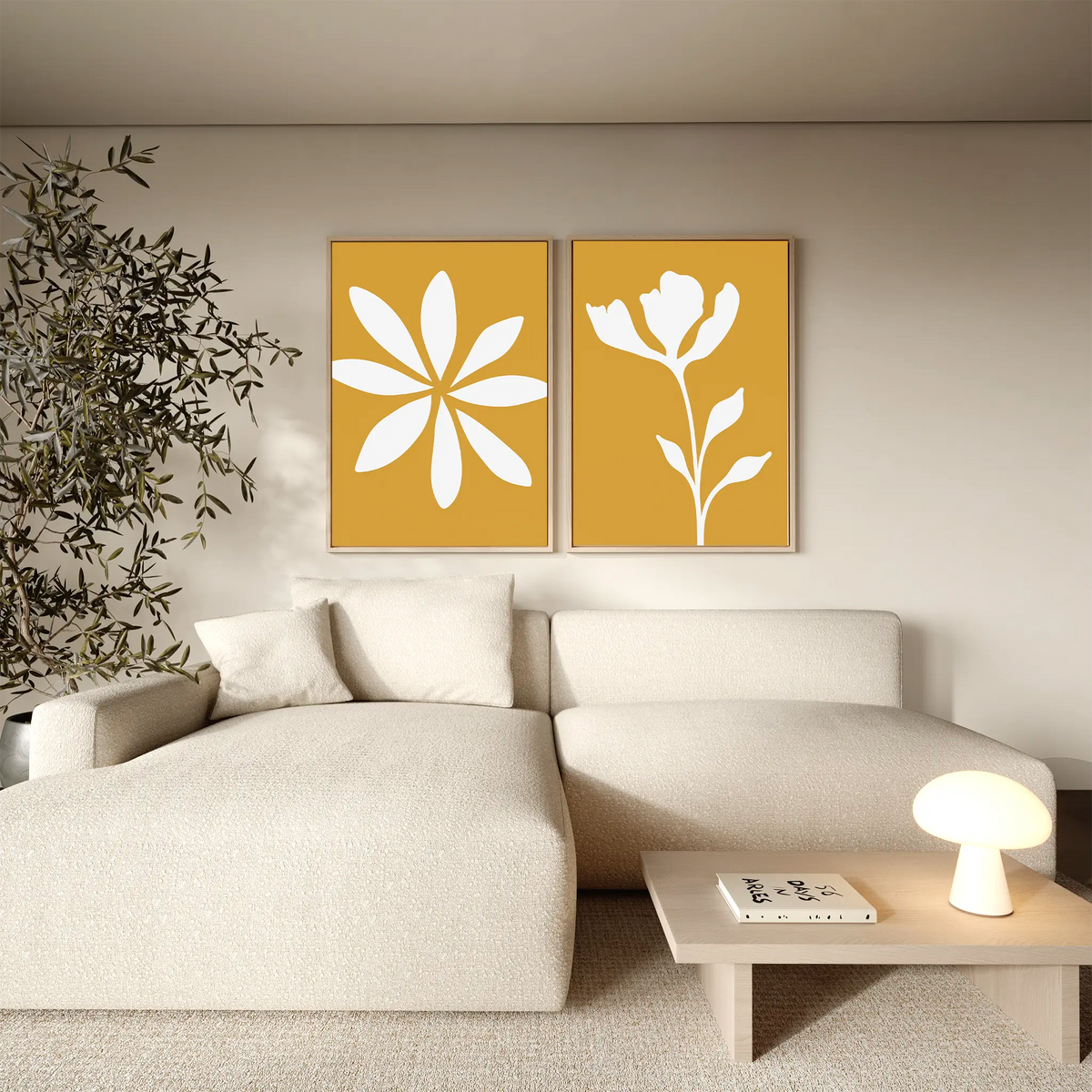 A modern living room features a cream sectional sofa, a small table with a round lamp and books, the Golden Bloom Set of 2 yellow floral wall art above the couch, and a leafy plant in the corner.