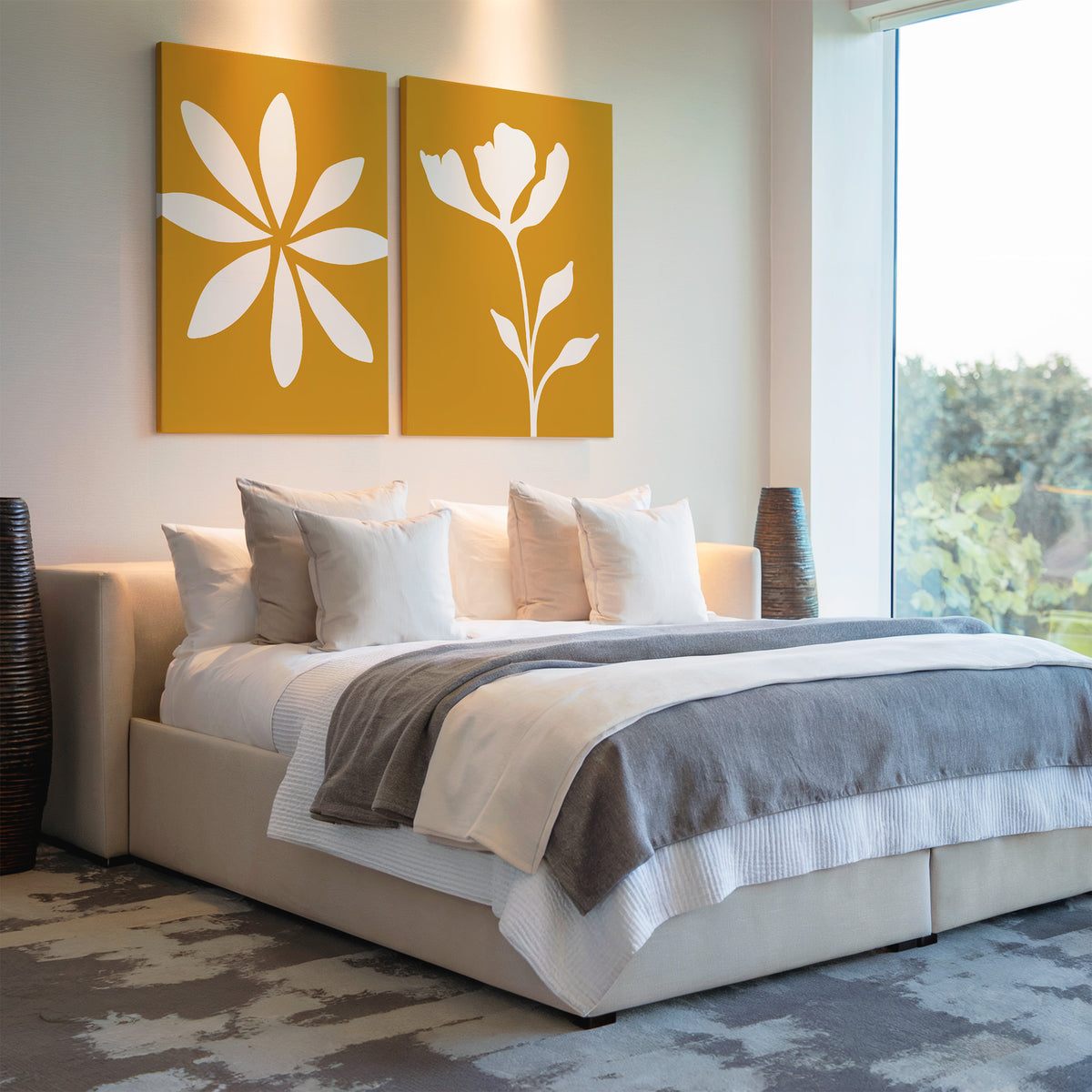 Modern bedroom with a large bed, gray and white bedding, several pillows, and the Golden Bloom Set of 2 floral wall art above the headboard. Natural light enters from a large window; two dark vases are beside the bed.