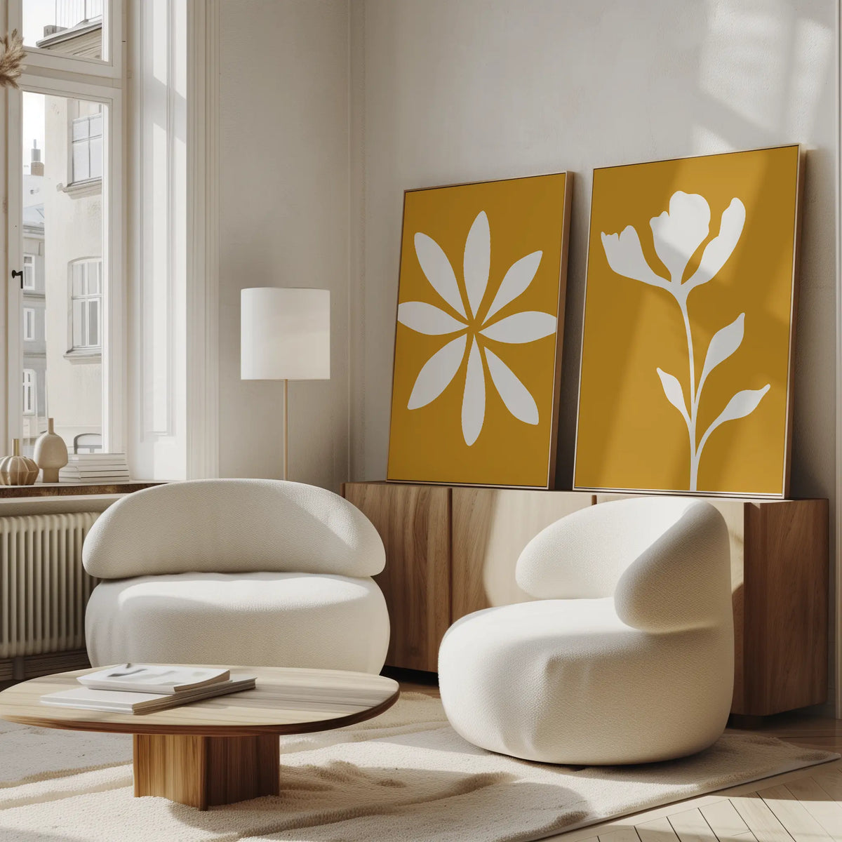 A modern living room features two rounded white chairs, a low wooden coffee table, and the Golden Bloom Set of 2 yellow wall art pieces with white floral designs. Large windows fill the neutral space with natural light.