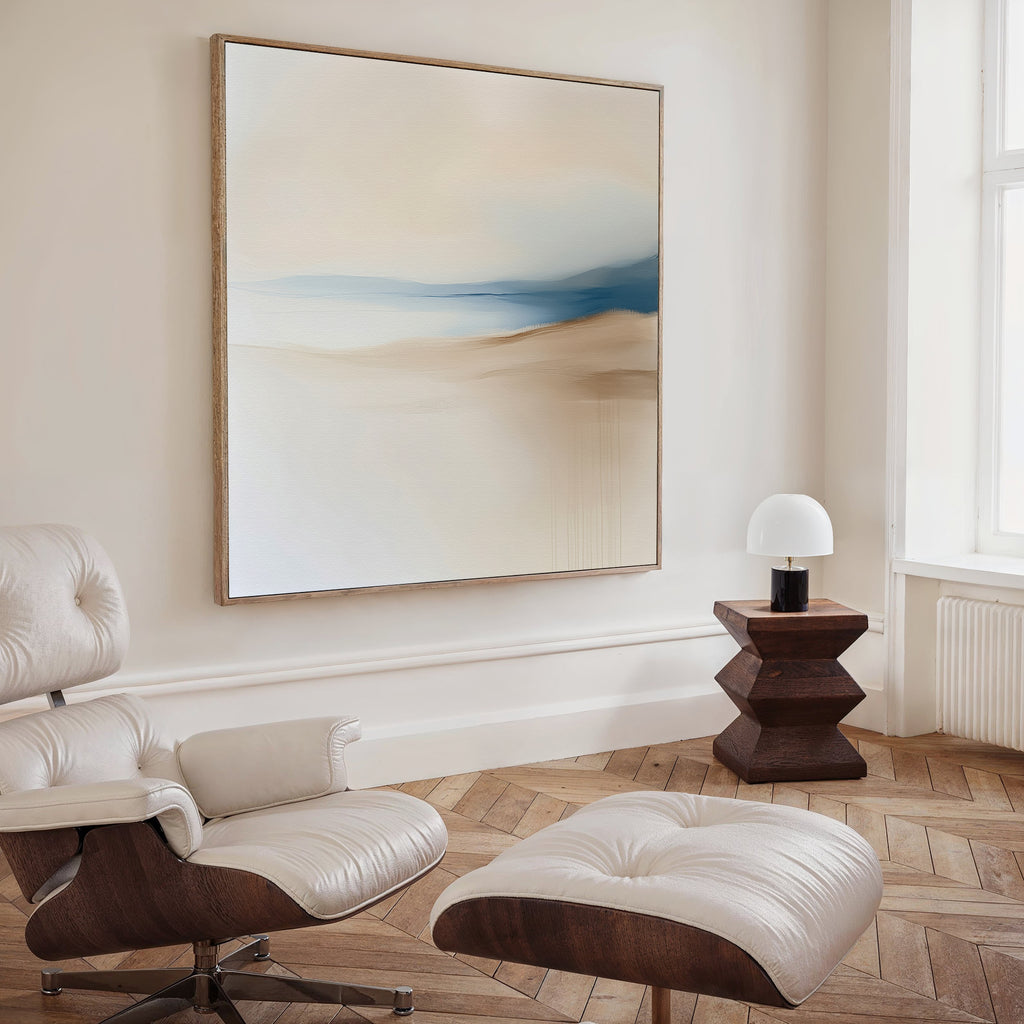 A modern living room features a cream lounge chair and ottoman, wood side table with white lamp, herringbone floor, and the Golden Drift Canvas Art in soft beige and blue tones for a refined touch.