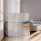 The Golden Drift Canvas Art, featuring soft beige and blue tones, rests against a white wall beside a modern white sofa and tall windows—ideal for adding contemporary style to your sunlit living room with parquet flooring.