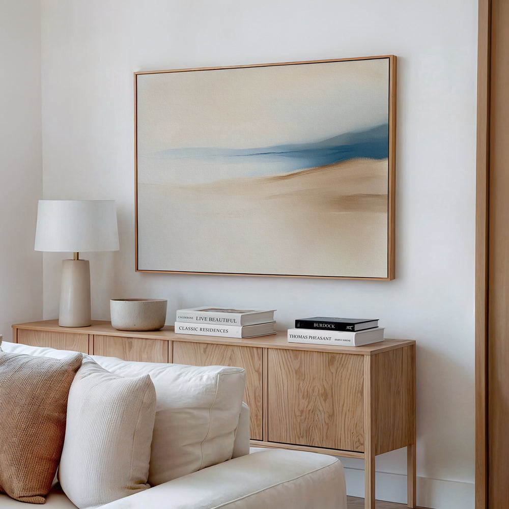 A minimal, modern living room featuring the Golden Drift Canvas Art – Horizontal in beige and blue tones above a cream sofa, with a light wooden sideboard, stacked books, a white lamp, and a bowl—perfect for inspiring your decor.