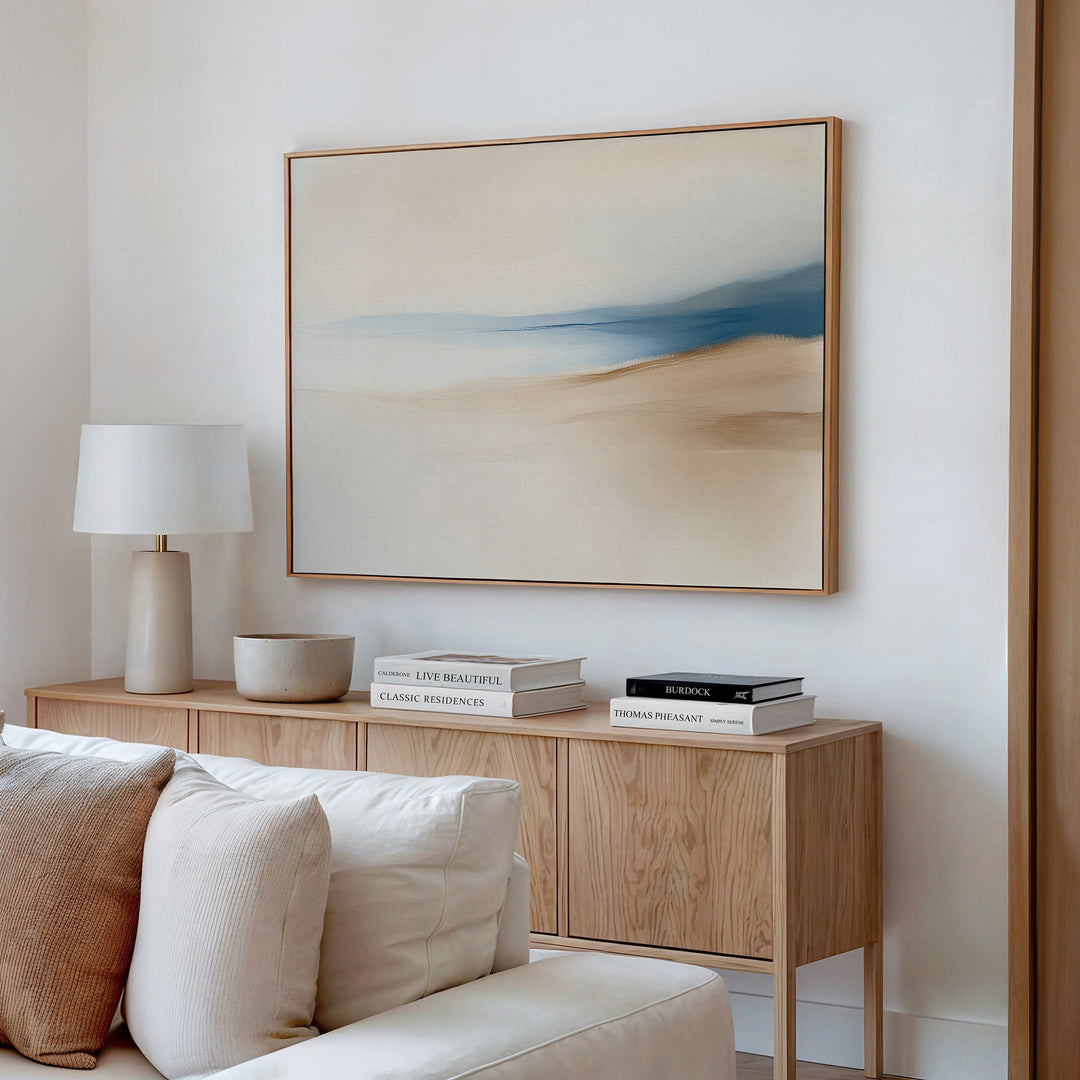 A minimal, modern living room featuring the Golden Drift Canvas Art – Horizontal in beige and blue tones above a cream sofa, with a light wooden sideboard, stacked books, a white lamp, and a bowl—perfect for inspiring your decor.
