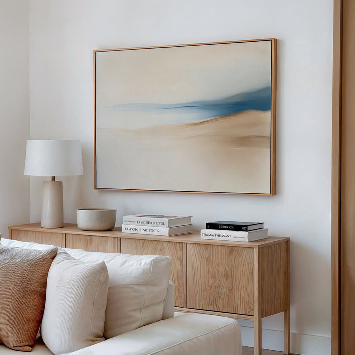 A minimal, modern living room featuring the Golden Drift Canvas Art – Horizontal in beige and blue tones above a cream sofa, with a light wooden sideboard, stacked books, a white lamp, and a bowl—perfect for inspiring your decor.