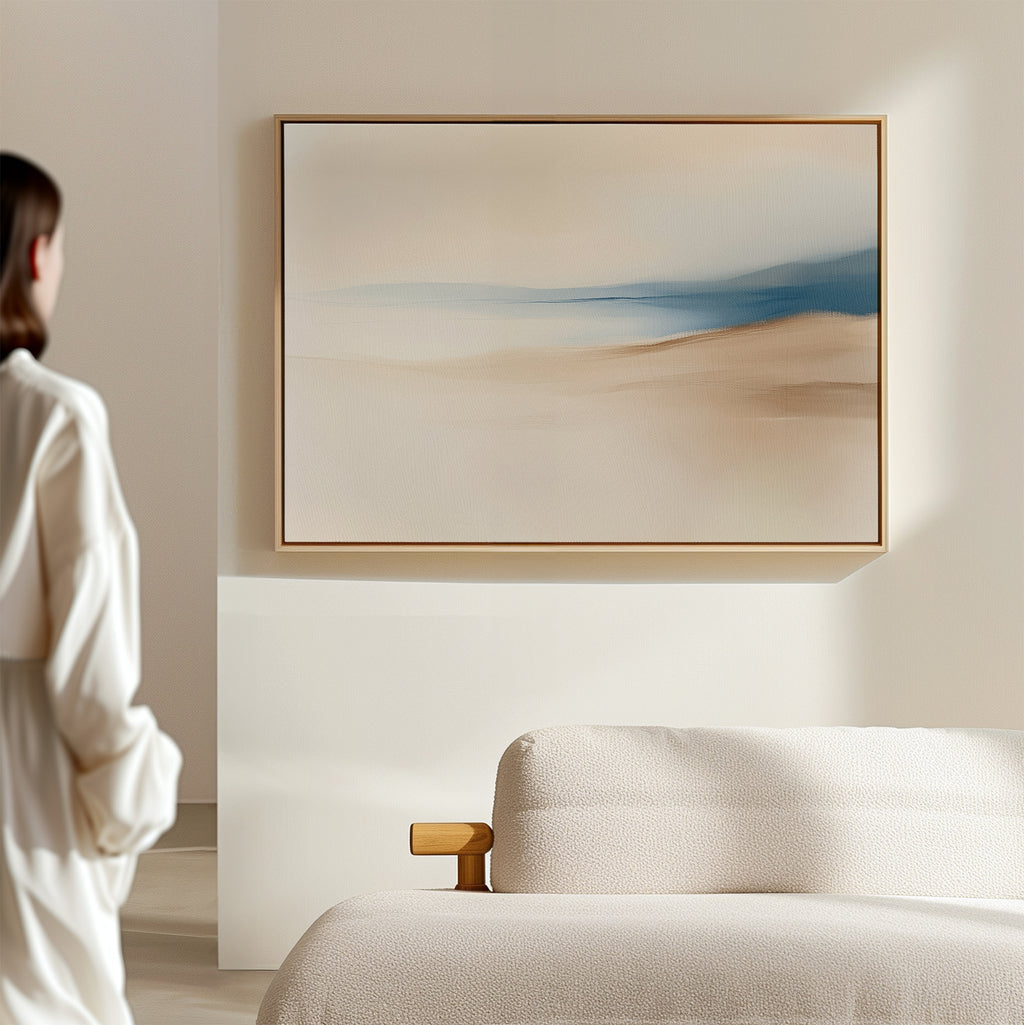 A person in a white outfit stands near a beige sofa, admiring the Golden Drift Canvas Art – Horizontal featuring soft beige and blue tones on a light-colored wall in a modern, minimalistic living room.