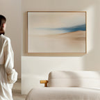 A person in a white outfit stands near a beige sofa, admiring the Golden Drift Canvas Art – Horizontal featuring soft beige and blue tones on a light-colored wall in a modern, minimalistic living room.