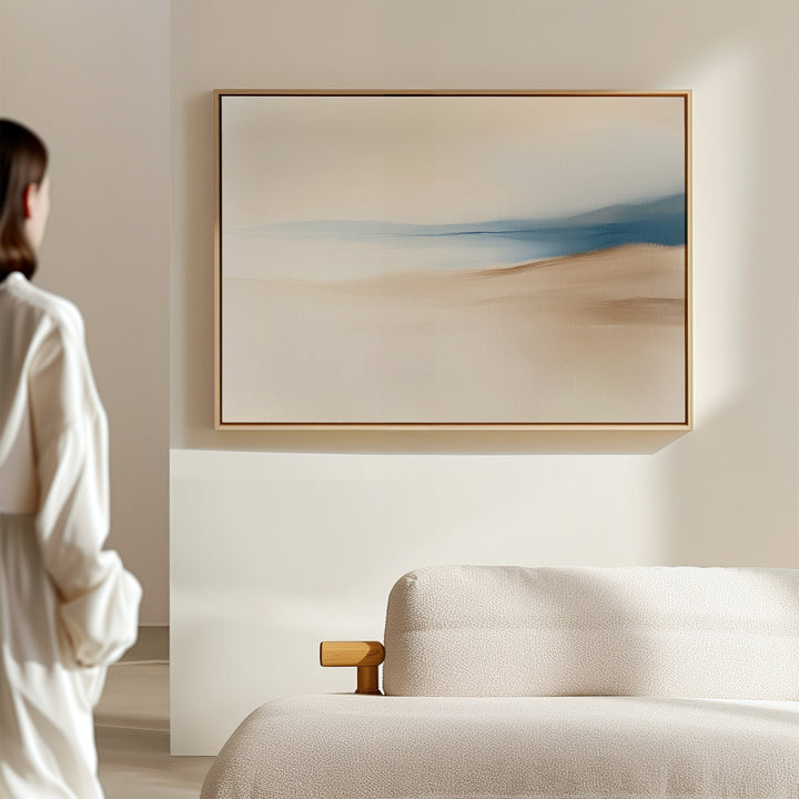 A person in a white outfit stands near a beige sofa, admiring the Golden Drift Canvas Art – Horizontal featuring soft beige and blue tones on a light-colored wall in a modern, minimalistic living room.