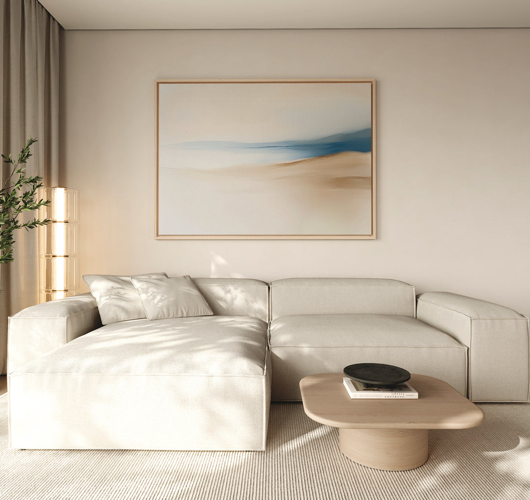 A modern living room features a light beige sectional, light wooden coffee table, black bowl, tall plant, and the Golden Drift Canvas Art – Horizontal with blue and beige tones. Soft natural light enhances the stylish decor.