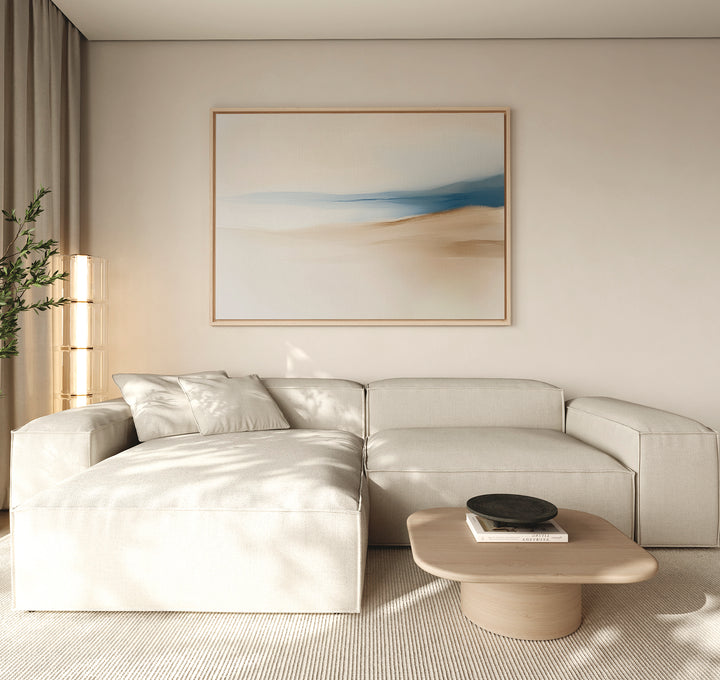 A modern living room features a light beige sectional, light wooden coffee table, black bowl, tall plant, and the Golden Drift Canvas Art – Horizontal with blue and beige tones. Soft natural light enhances the stylish decor.