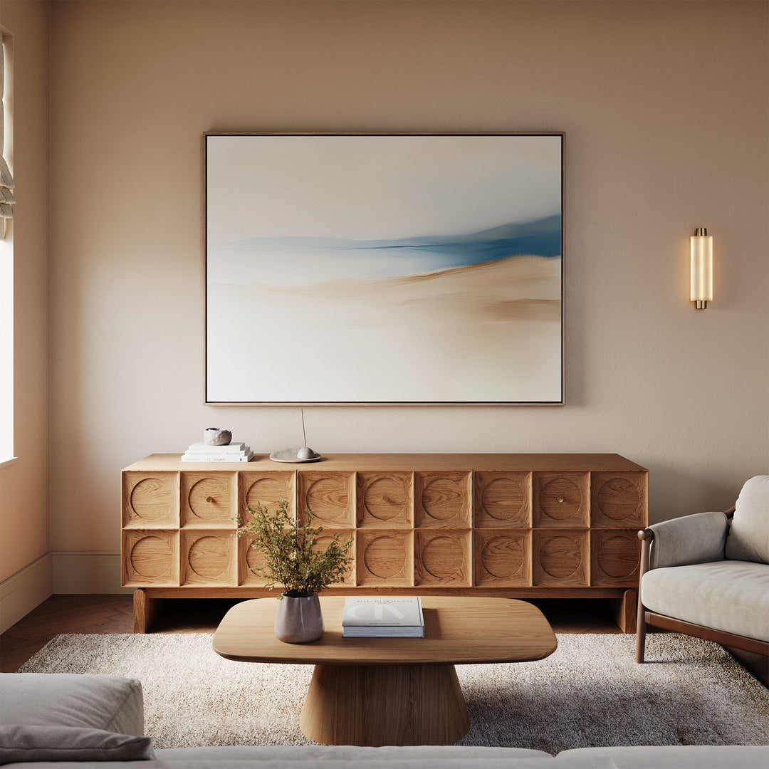 A modern living room with light beige walls features a wooden sideboard, minimalist coffee table, and a beige sofa. The Golden Drift Canvas Art – Horizontal hangs above, elevating the space. A vase with flowers decorates the table.
