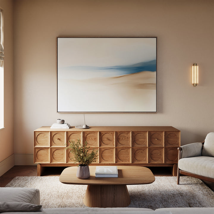 A modern living room with light beige walls features a wooden sideboard, minimalist coffee table, and a beige sofa. The Golden Drift Canvas Art – Horizontal hangs above, elevating the space. A vase with flowers decorates the table.