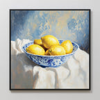 The Golden Fruit Canvas Art features a blue and white patterned bowl of lemons on a white cloth with a soft blue and yellow background. Perfect for vintage home decor, this print comes framed in sleek black.