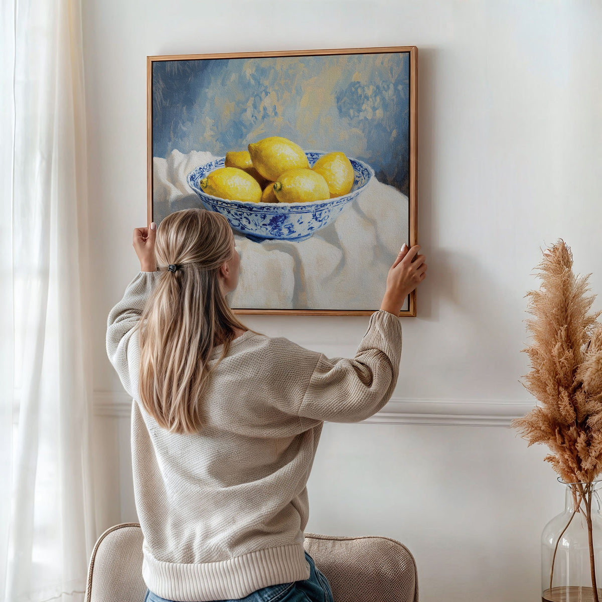 A woman with a blonde ponytail hangs the Golden Fruit Canvas Art—a print of lemons in a blue and white bowl—on a white wall, as pampas grass decor and soft natural light add vintage charm to the cozy room.