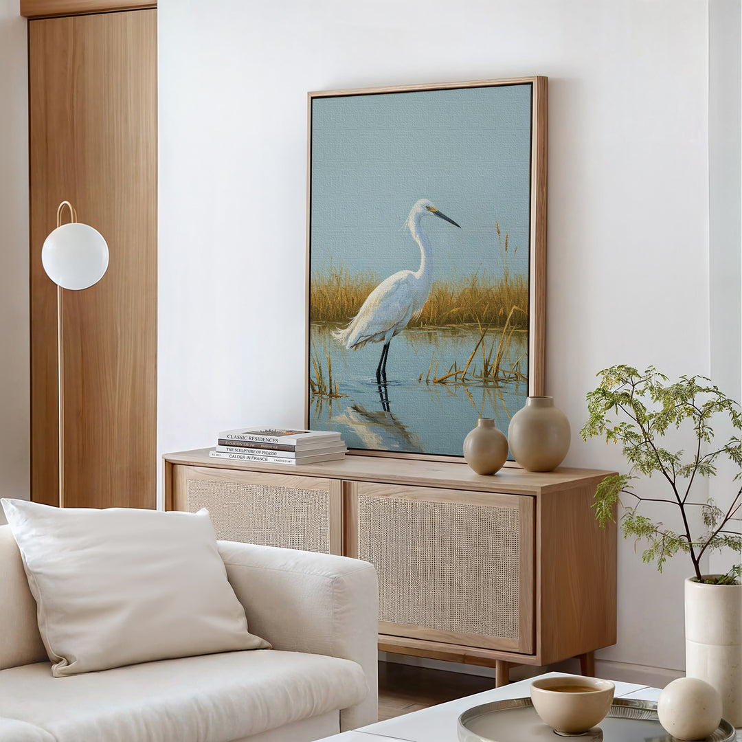 A modern living room with neutral tones features Golden Marsh Egret Canvas Art as elegant wall decor, complemented by a white sofa, serene accents, a leafy corner plant, and a sideboard adorned with vases and books.