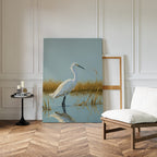 The Golden Marsh Egret Canvas Art, featuring a white heron in water, rests against a white wall. Modern accents and a cushioned chair on herringbone wood flooring enhance the serene home decor.