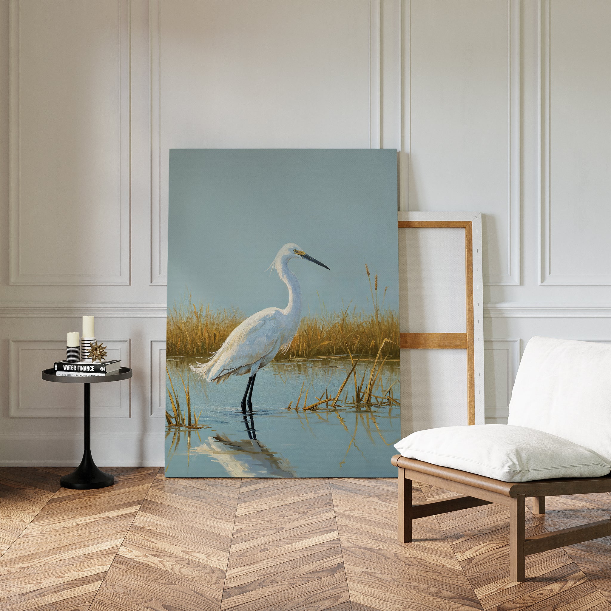 The Golden Marsh Egret Canvas Art, featuring a white heron in water, rests against a white wall. Modern accents and a cushioned chair on herringbone wood flooring enhance the serene home decor.