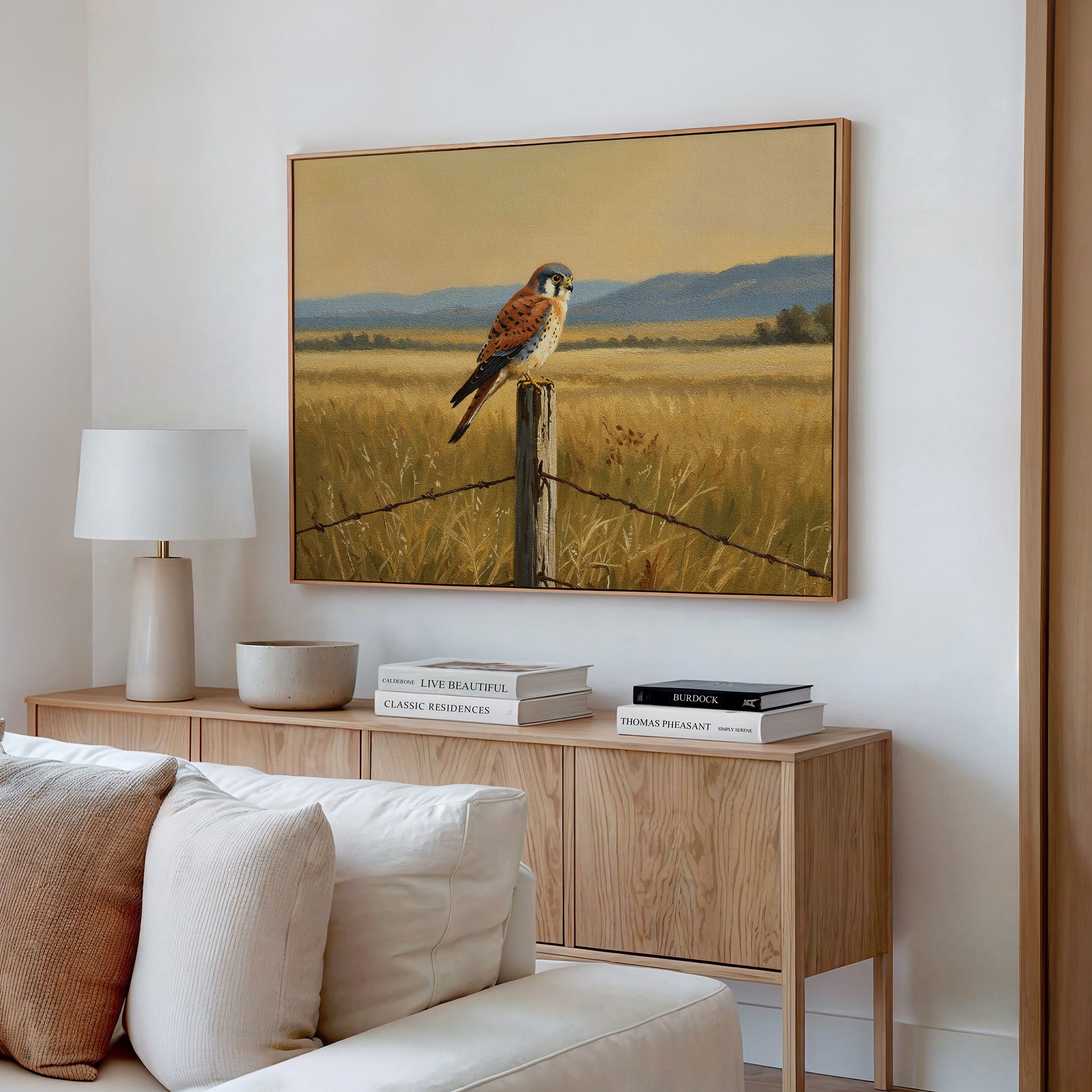 A modern living room with a light wood sideboard, white lamp, stacked books, and nature-inspired wall decor featuring the Golden Plains Canvas Art—a painting of a bird on a fence post in a golden field with mountains in the background.