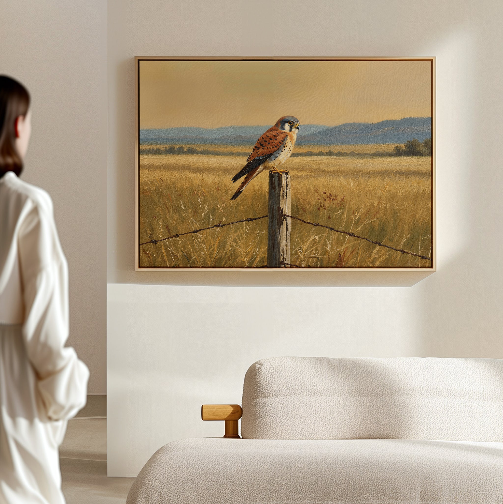 A woman in a white robe stands in a minimalist room, admiring the Golden Plains Canvas Art—a framed painting of a kestrel perched on a fence post in a golden field—bringing striking nature-inspired decor to the serene space.