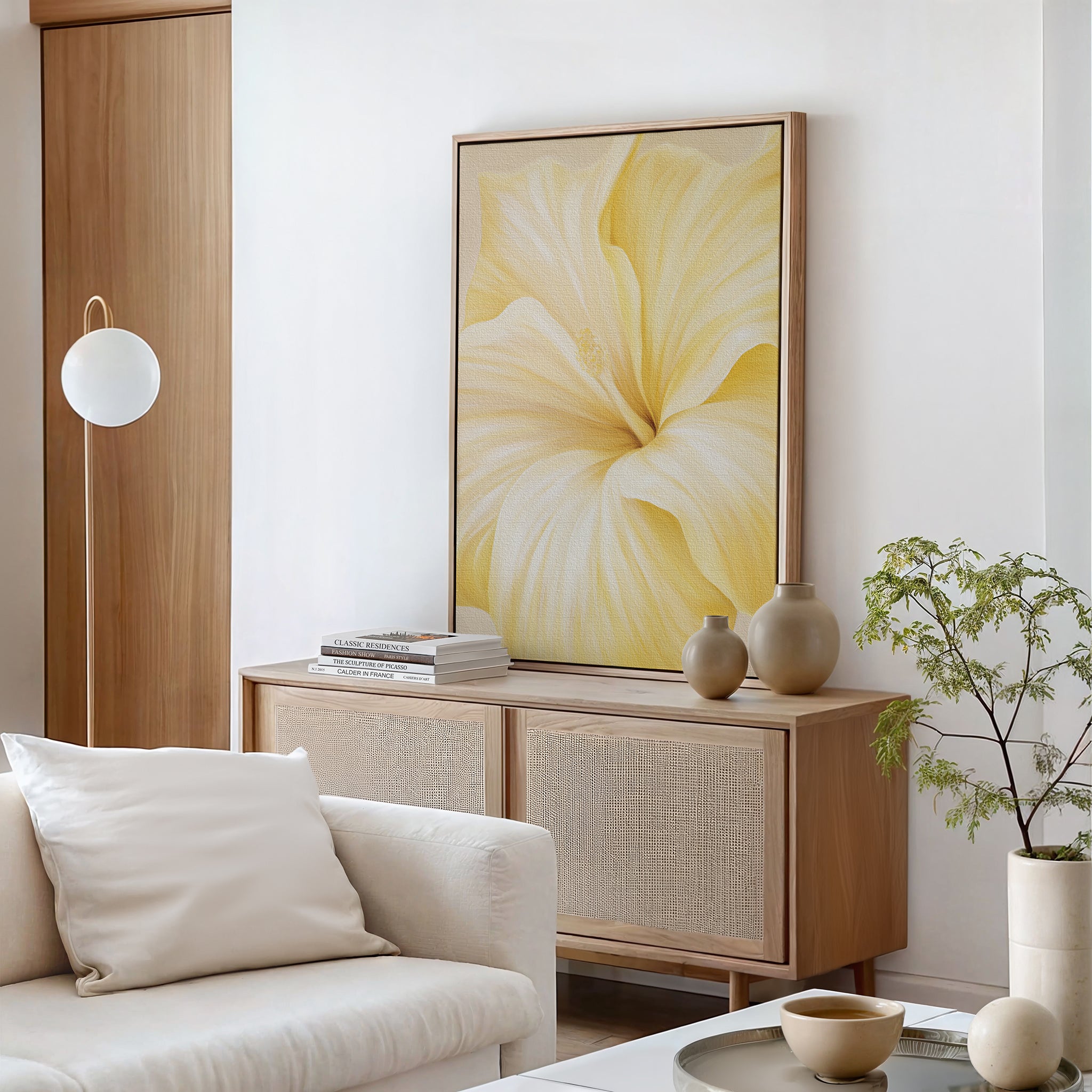 A modern living room with a light wood cabinet, the Golden Radiance Canvas Art featuring yellow flowers, neutral decor, a white sofa, stacked books, ceramic vases, and a small potted plant against crisp white walls.