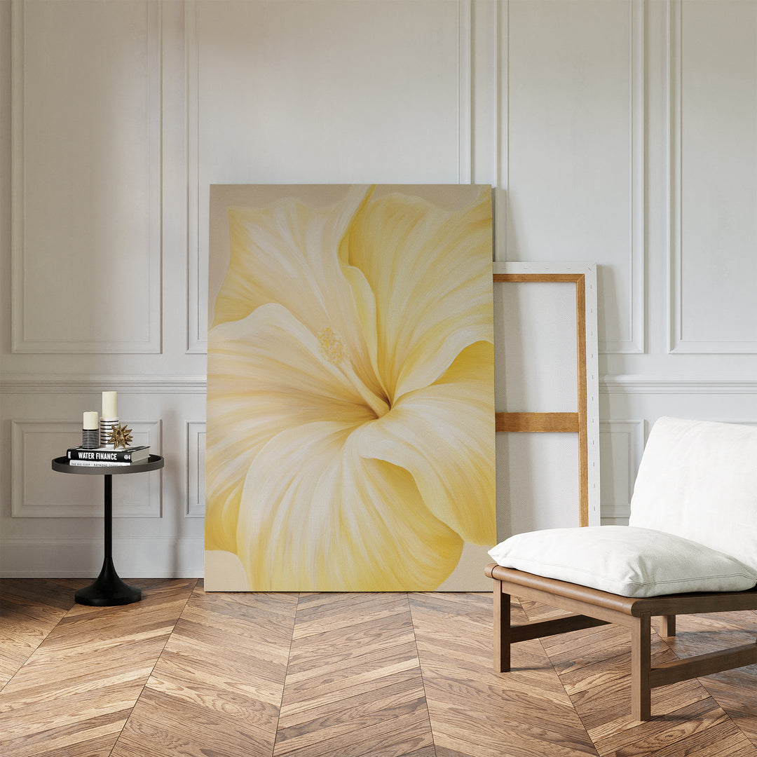 The Golden Radiance Canvas Art, with its close-up yellow flower design, leans against a white paneled wall, bringing striking minimalist style to the room alongside a small round table with candles and a padded wooden chair on herringbone floors.
