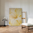 The Golden Radiance Canvas Art, with its close-up yellow flower design, leans against a white paneled wall, bringing striking minimalist style to the room alongside a small round table with candles and a padded wooden chair on herringbone floors.
