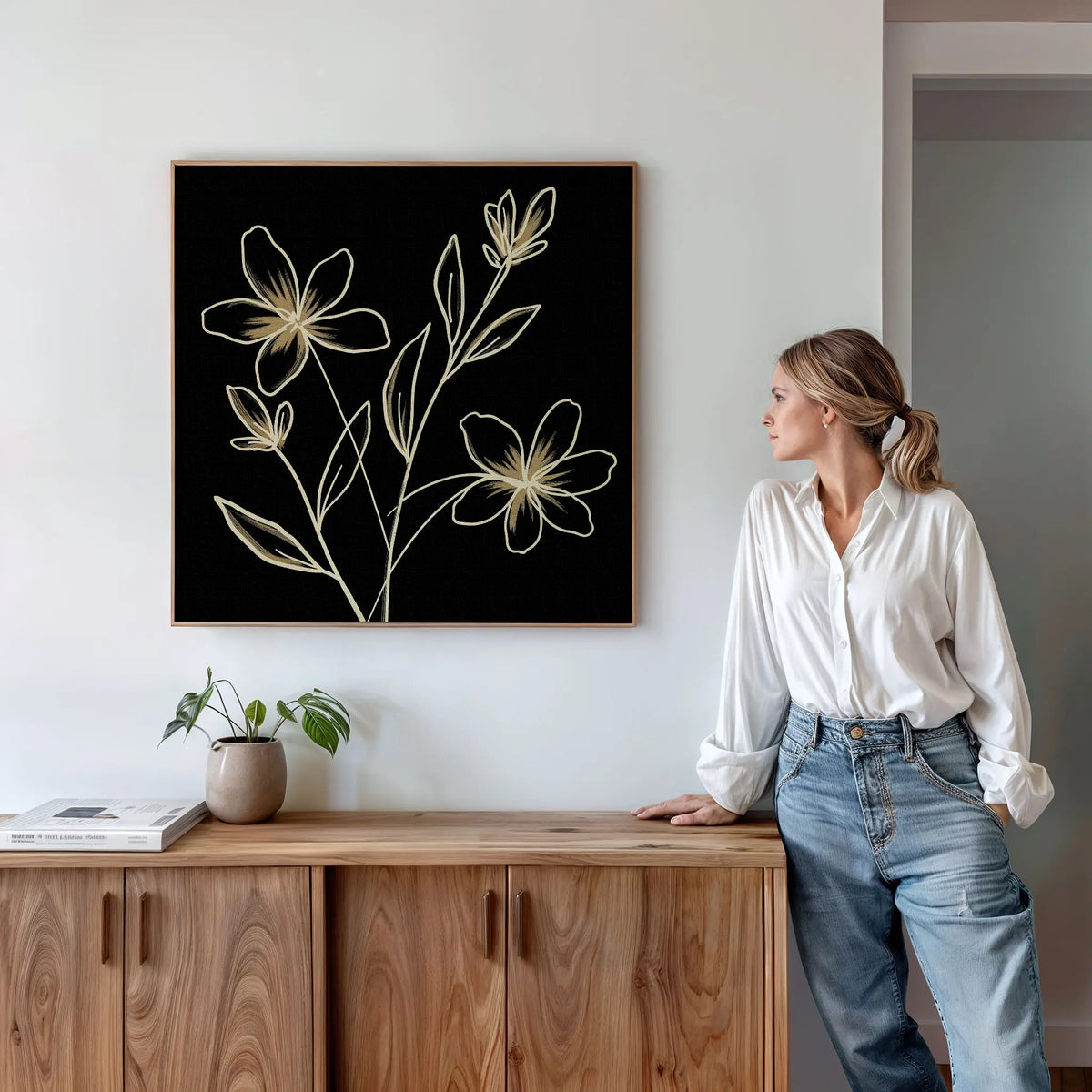 A woman in a white blouse and jeans leans against a wooden cabinet, admiring the Golden Stems Canvas Art—a minimalist piece with light floral outlines. A small plant and books accent the modern, bright room’s elevated decor.