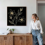 A woman in a white blouse and jeans leans against a wooden cabinet, admiring the Golden Stems Canvas Art—a minimalist piece with light floral outlines. A small plant and books accent the modern, bright room’s elevated decor.