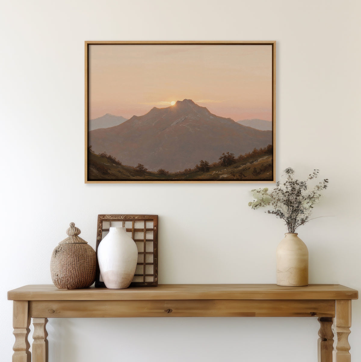 A wooden table with neutral-toned vases and decor sits against a white wall, topped by the Golden Summit Canvas Art depicting a mountain sunset. Dried flowers and a woven basket complete this inviting living room scene.