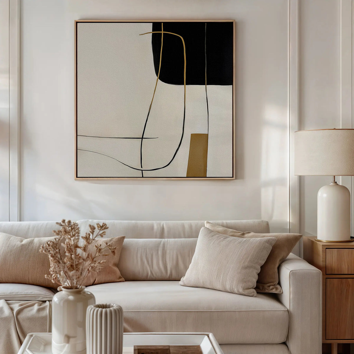 A modern living room with a beige sofa, light cushions, minimalist decor, a side table and lamp, dried flowers in a vase, and the Golden Thread Canvas Art featuring black, beige, and gold lines displayed on the wall.