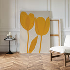 The Golden Tulips Abstract Canvas Art leans against a white paneled wall beside a wooden chair with a white cushion and a black side table with candles, creating stylish living room decor on a herringbone wood floor.