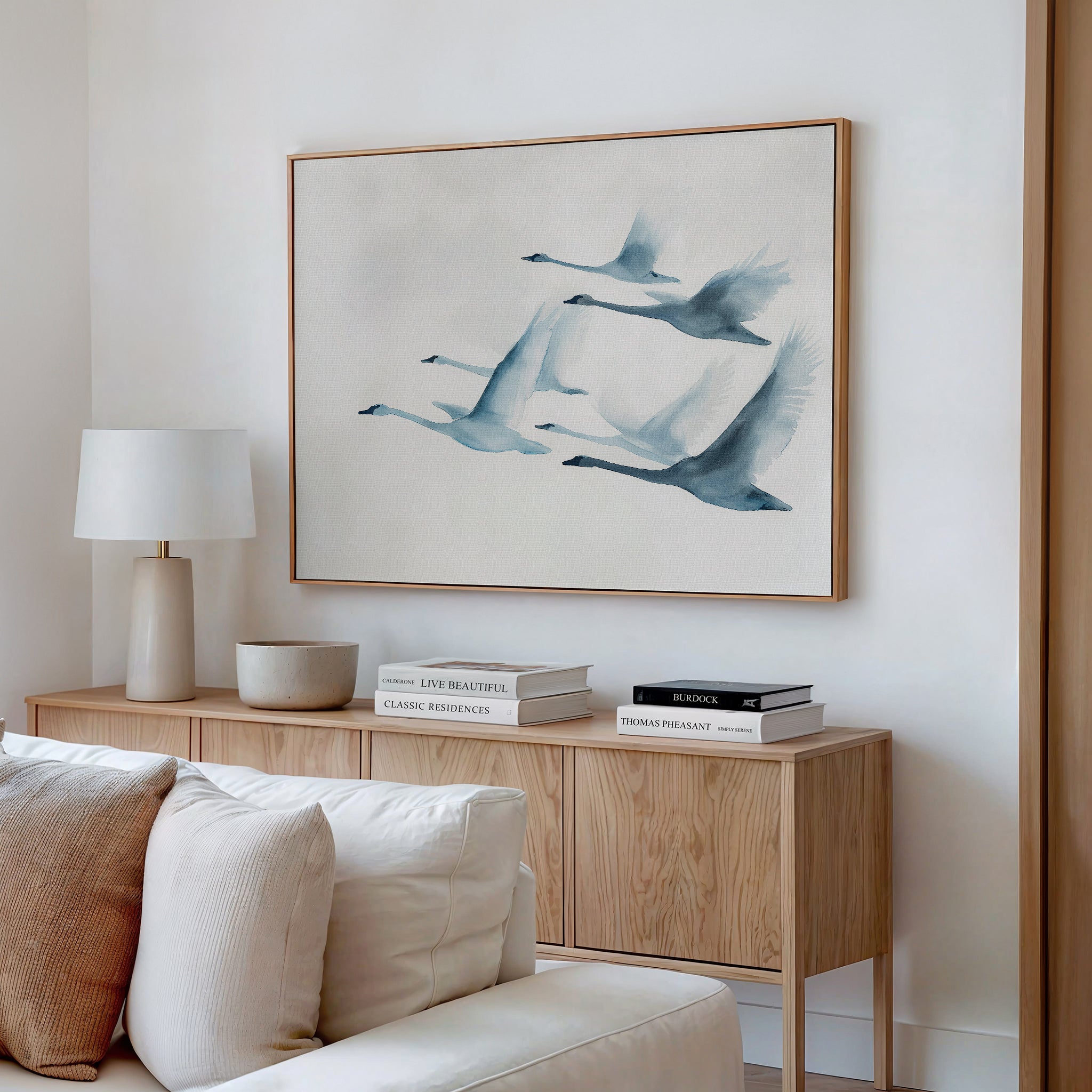 A minimalist living room features a wooden sideboard with books, a lamp, and decor beneath the Graceful Flock Canvas Art—a framed painting of five blue-toned birds. A cream sofa appears in the foreground, complementing this modern nature decor.