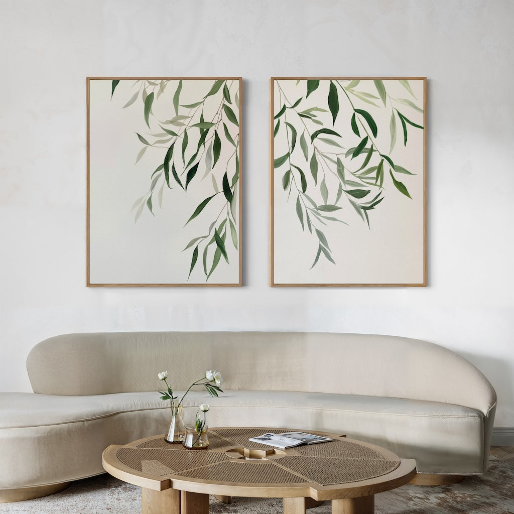 A minimalist living room features a curved beige sofa, a round wooden coffee table with vases and flowers, and the Graceful Leaves Set of 2 wall art prints showcasing elegant green foliage decor.