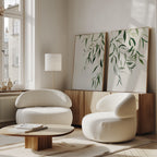 A modern living room with two white, rounded armchairs, a low wooden coffee table, and the Graceful Leaves Set of 2 wall art. Tall windows fill the minimal space with natural light, adding to its cozy decor.