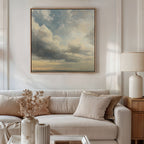 A cozy living room features a beige sofa, neutral cushions, vintage decor, a coffee table with dried flowers and a lamp, and the Heavenly Drift Canvas Art—a large cloudy sky print—above the sofa.